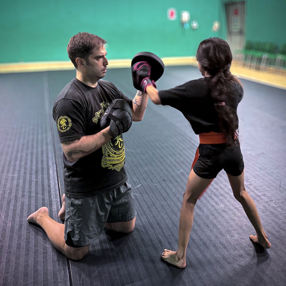 Paul kneeling to hold pads at the right height while coaching a trainee's hook
